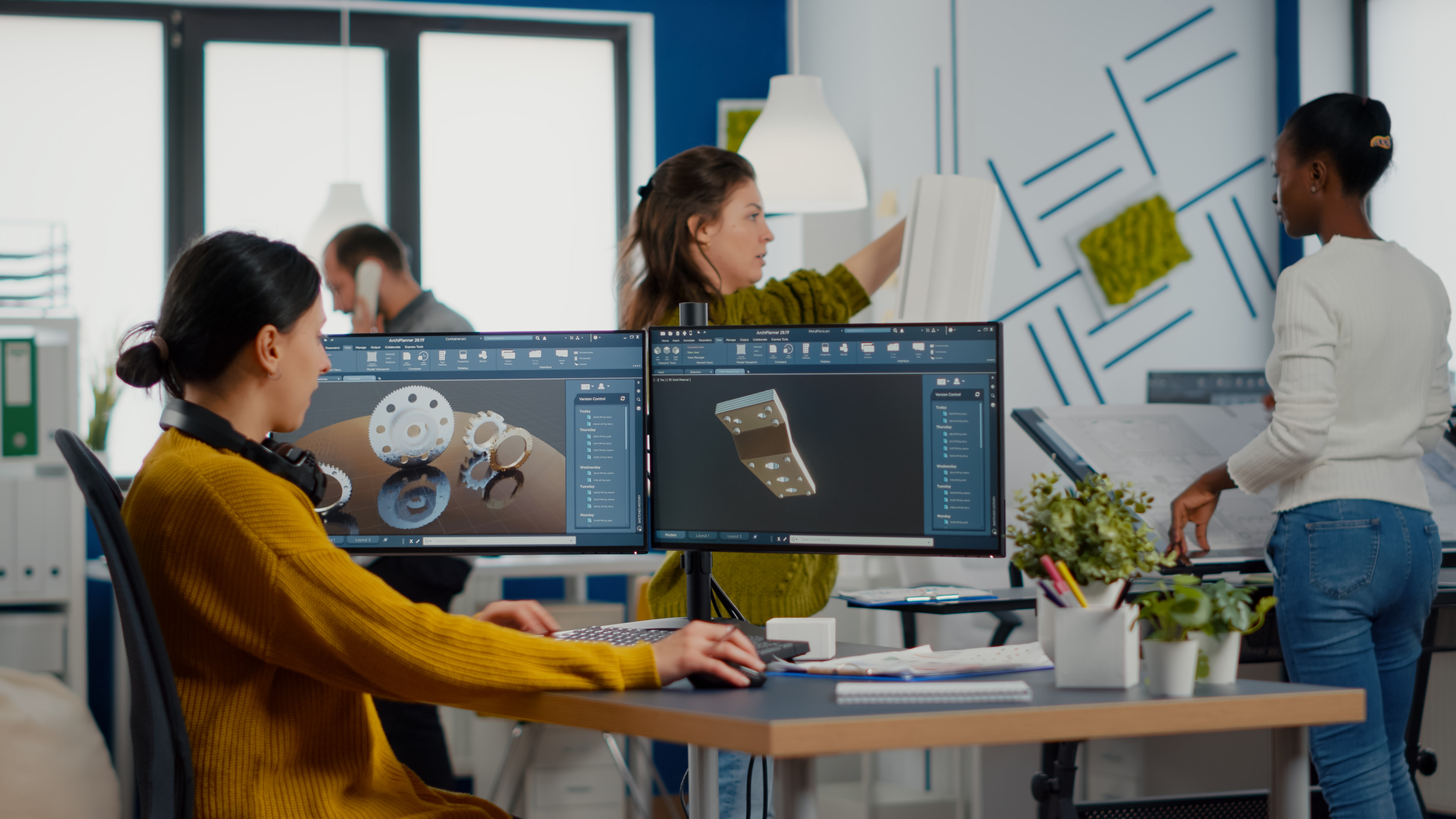 Person mit Kopfh&ouml;rern um den Hals sitz an Schreibtisch vor zwei gro&szlig;en Monitoren auf denen Renderings von Zahnr&auml;dern und Bauteilen zu sehen sind, im Hintergrund weitere Personen stehend in B&uuml;roraum