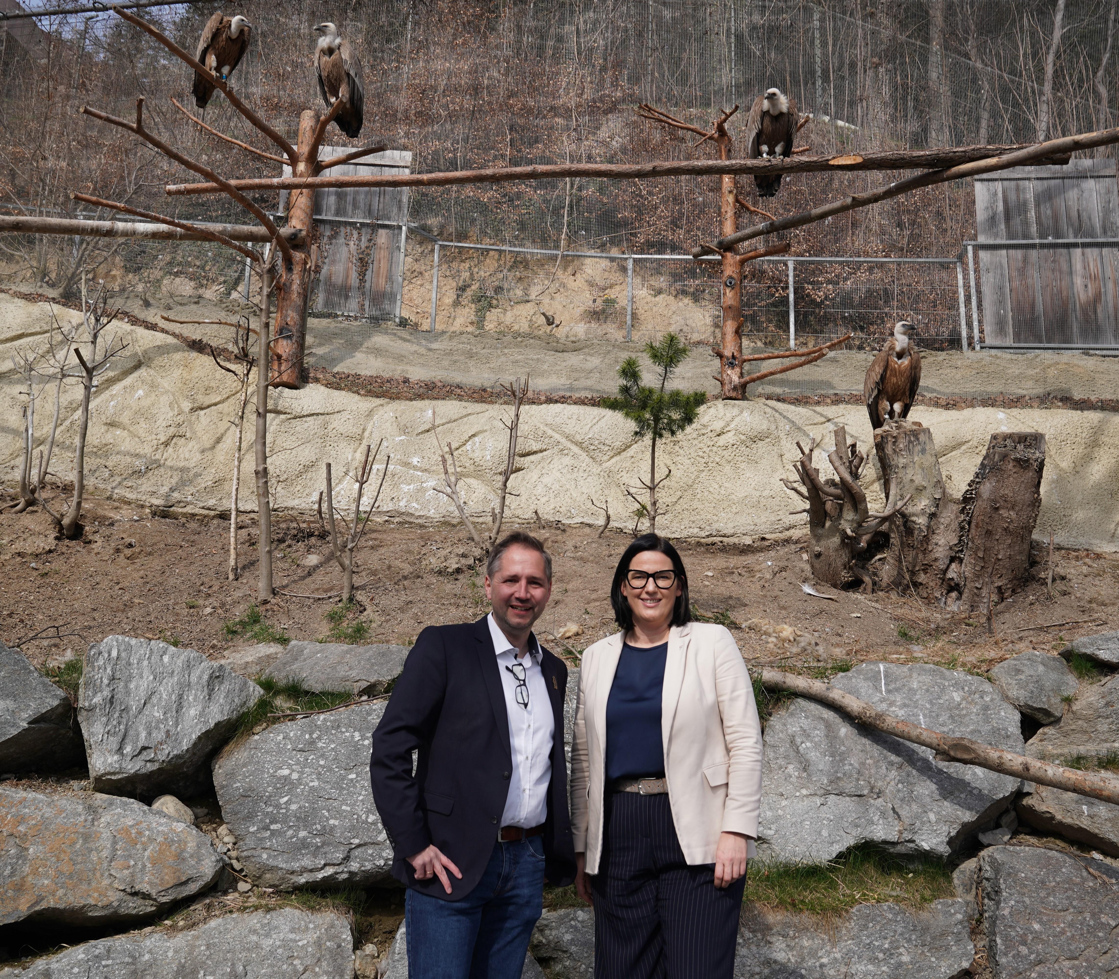 Zwei Erwachsene stehen vor einem gro&szlig;en Gehege mit mehreren Geiern, die auf Holzst&auml;mmen sitzen. Die Personen sind gepflegt gekleidet und l&auml;cheln in die Kamera. Im Hintergrund sind Felsen, B&auml;ume und ein abgesperrter Naturbereich zu sehen.
