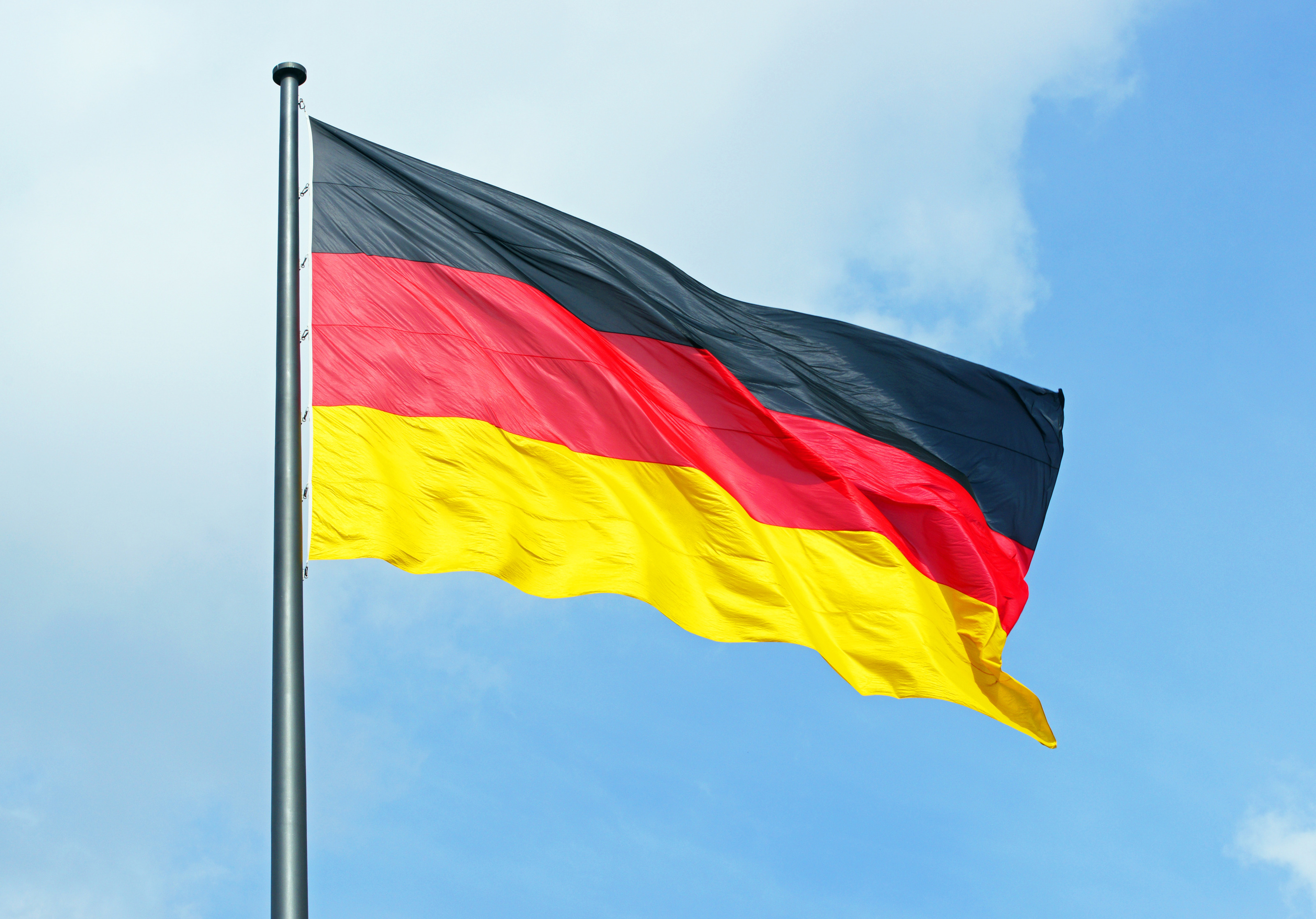 An einer Metallstange ist die Flagge Deutschlands montiert. Die Flagge weht im Wind. Dahinter ist der blaue Himmel mit einigen wei&szlig;en Wolken