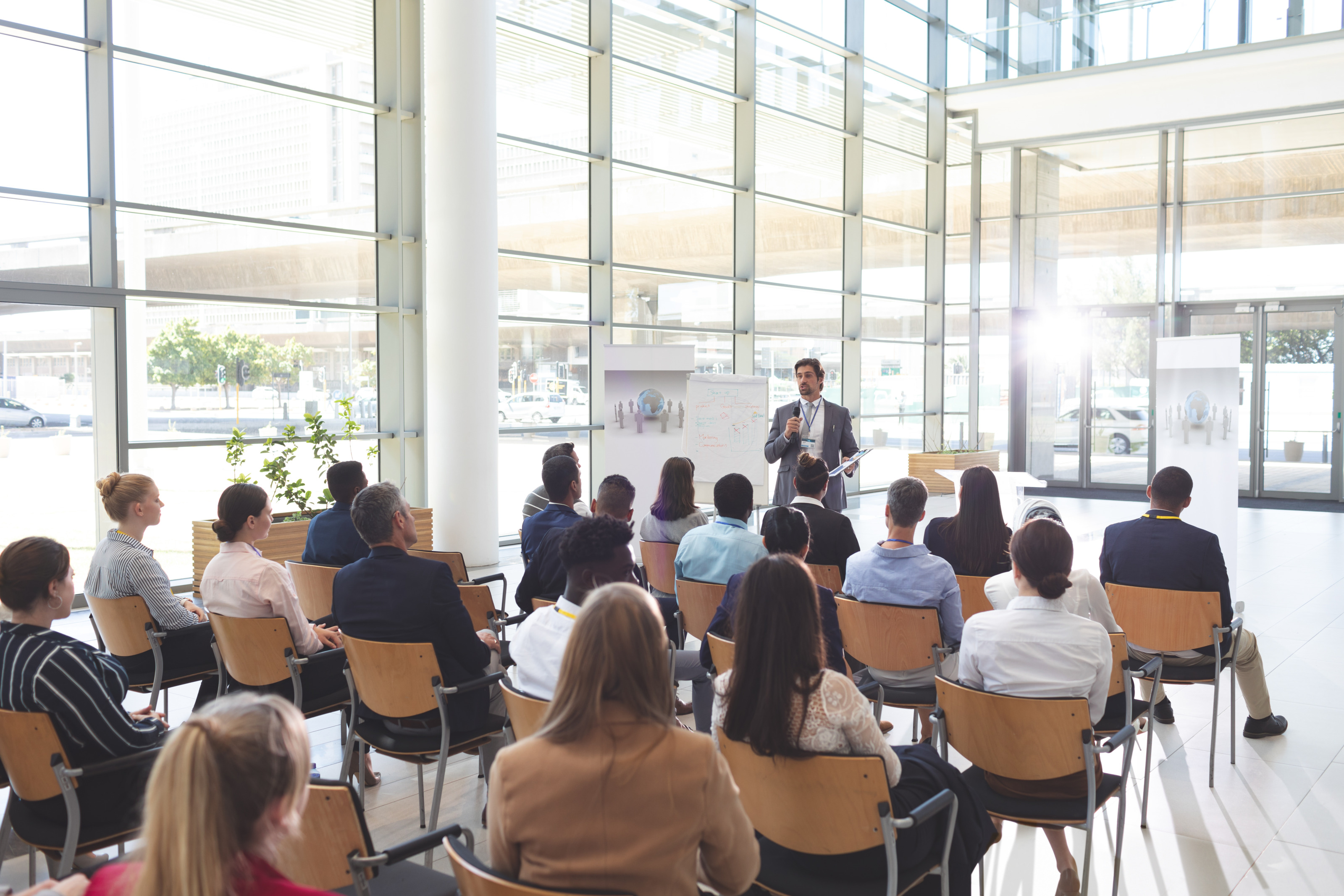 Vortragende Person steht in einem hellen, gl&auml;sernen Raum vor Publikum
