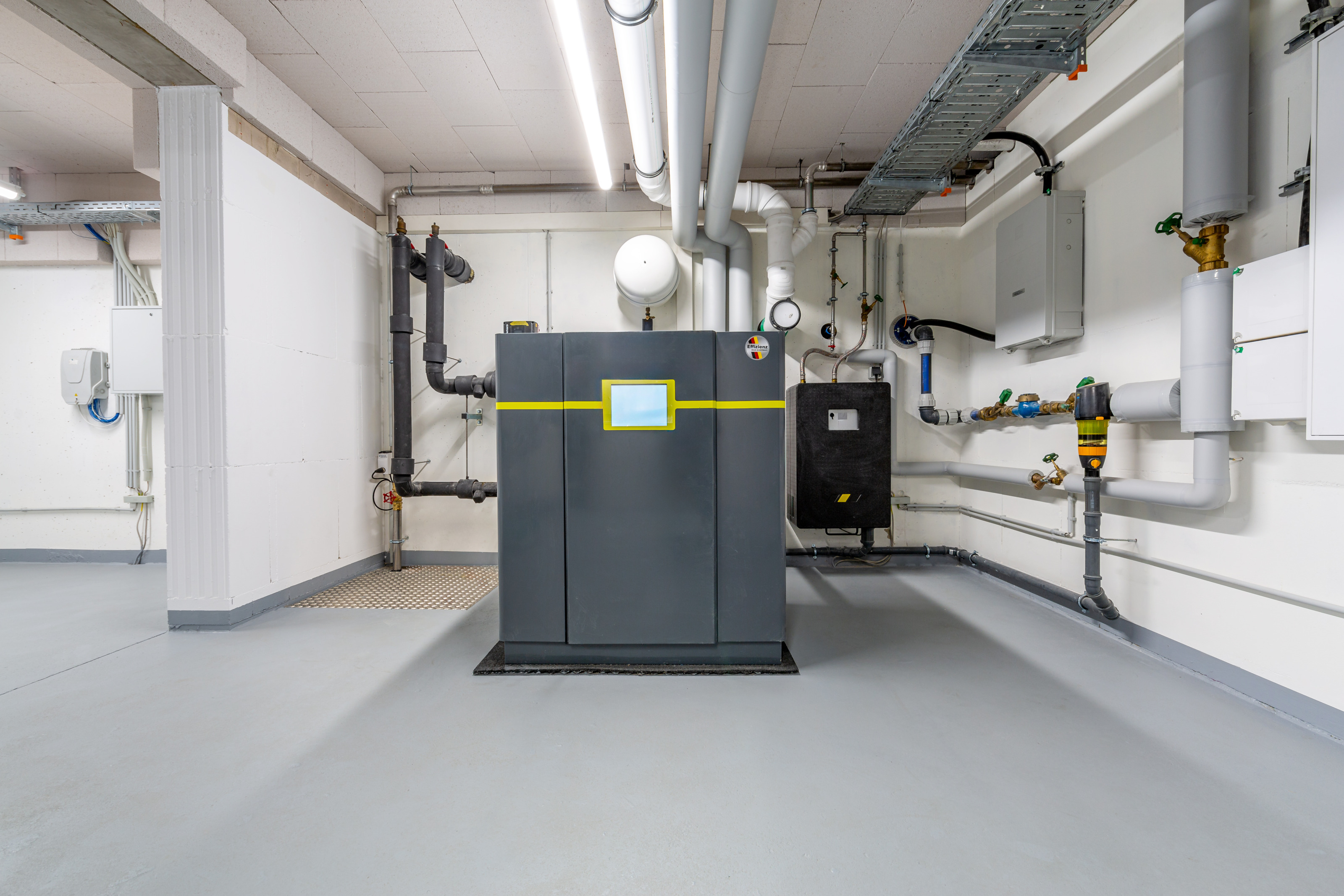 Ansicht einer W&auml;rmepumpe: Dunkelgrauer metallischer Block mit gelber Linie und mittig platziertem Display in wei&szlig;em Keller, ringsum Leitungen und Rohre verlaufend
