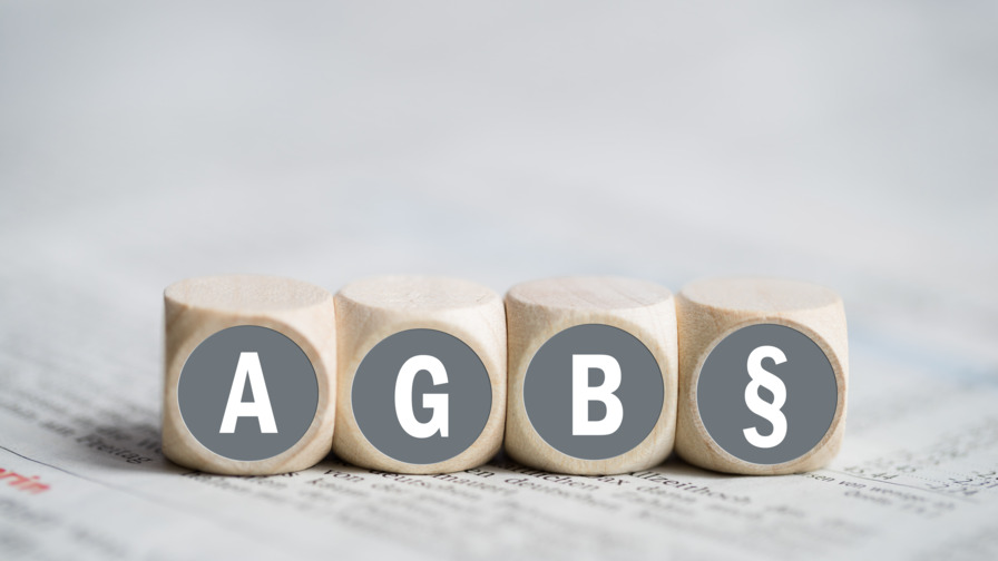 Holzwürfel mit Aufschrift AGB und Paragraphen-Symbol in weißer Schrift mit grau hinterlegtem Kreis, in der Unschärfe zeigt sich darunter eine ausgebreitete Tageszeitung