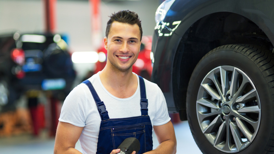 Eine Person steht mit Werkzeug in der Hand neben einem Auto auf einer Hebebühne, um die Reifen zu wechseln und lächelt in die Kamera.