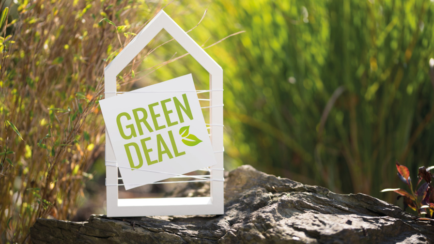 Weiße Form eines Häuschen mit Schild mit grüner Schrift und Symbol eines Blattes Green Deal auf Stein platziert, im Hintergrund verschwommen grünbraun Schilf