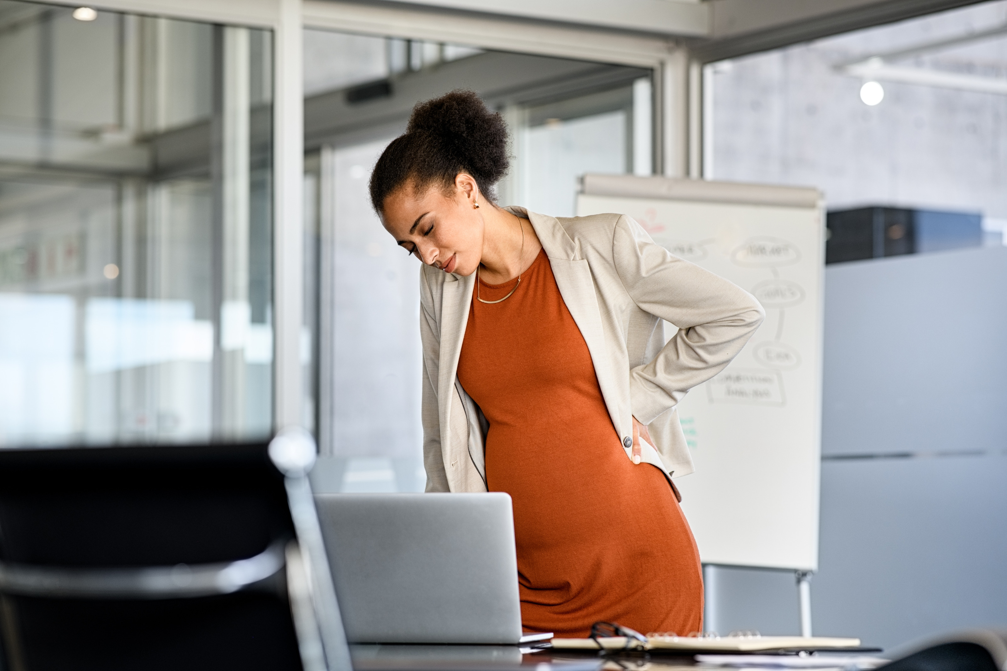 Eine schwangere Person steht in einem Büro und bückt sich über den Schreibtisch. 