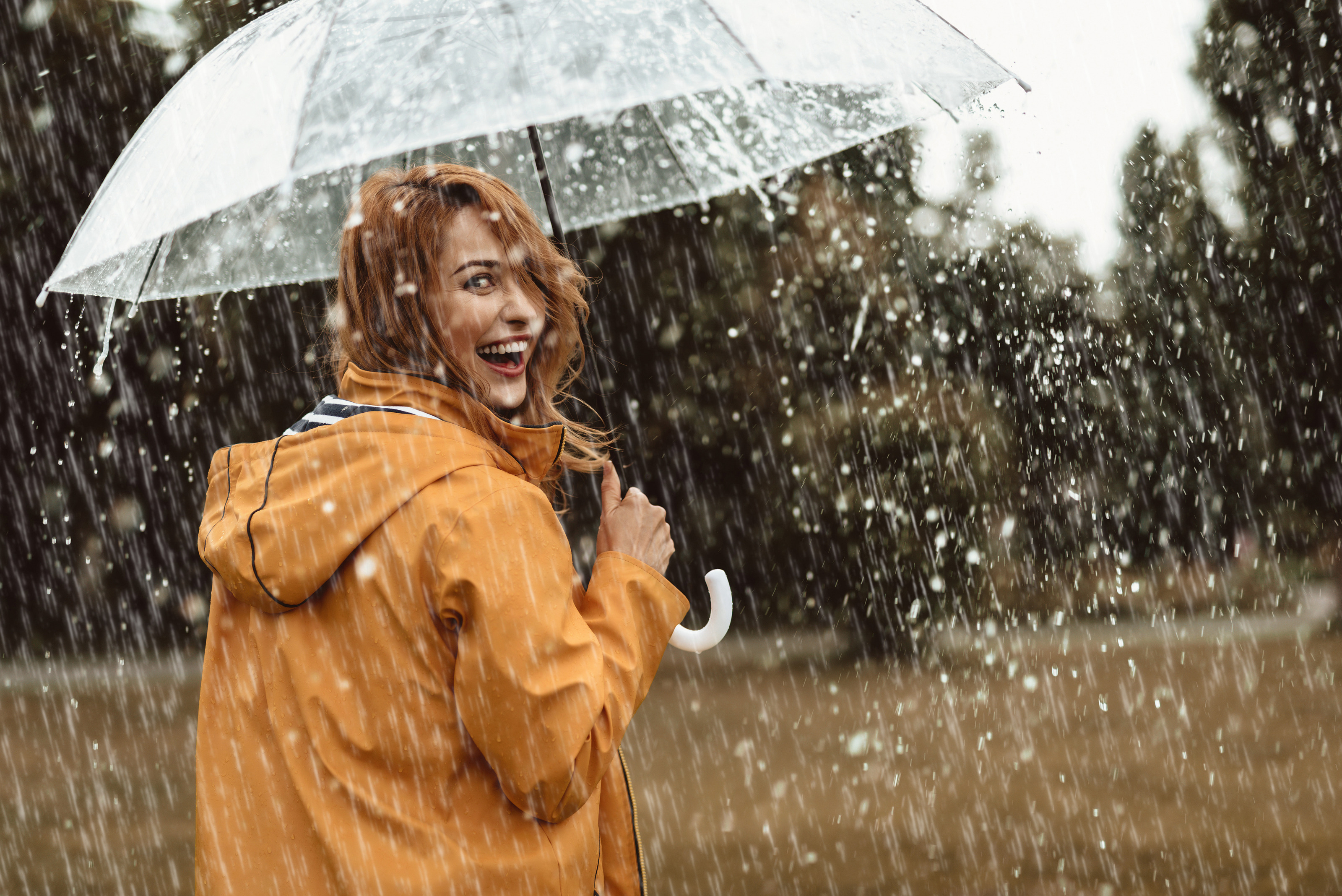 R&uuml;ckenansicht einer Person, die sich zur Kamera umdreht und lacht. Die tr&auml;gt eine orange Regenjacke und h&auml;lt einen Regenschirm. Die Person steht auf einer Wiese. Im Hintergrund sin gr&uuml;ne B&auml;ume. Es regnet