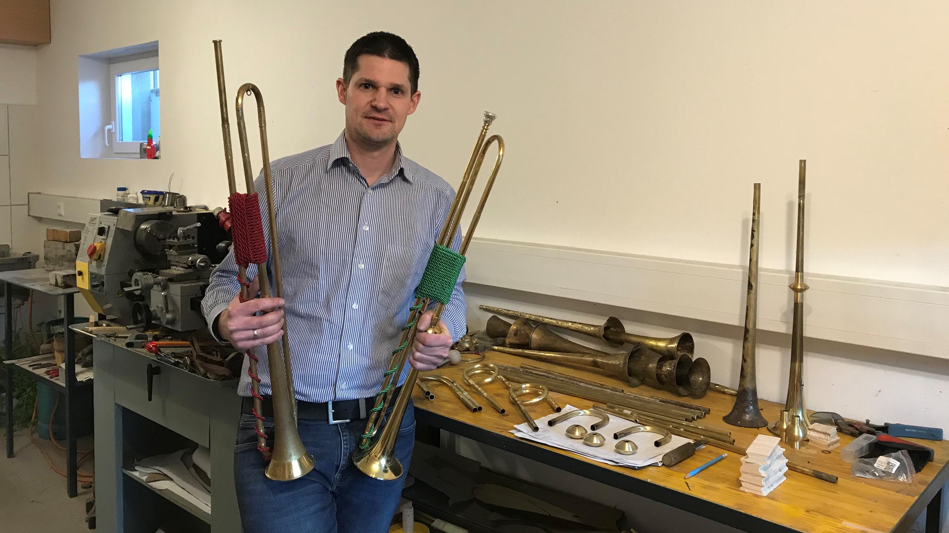 Harald Schuh steht mit zwei Barocktrompeten in den H&auml;nden in einer Werkstatt