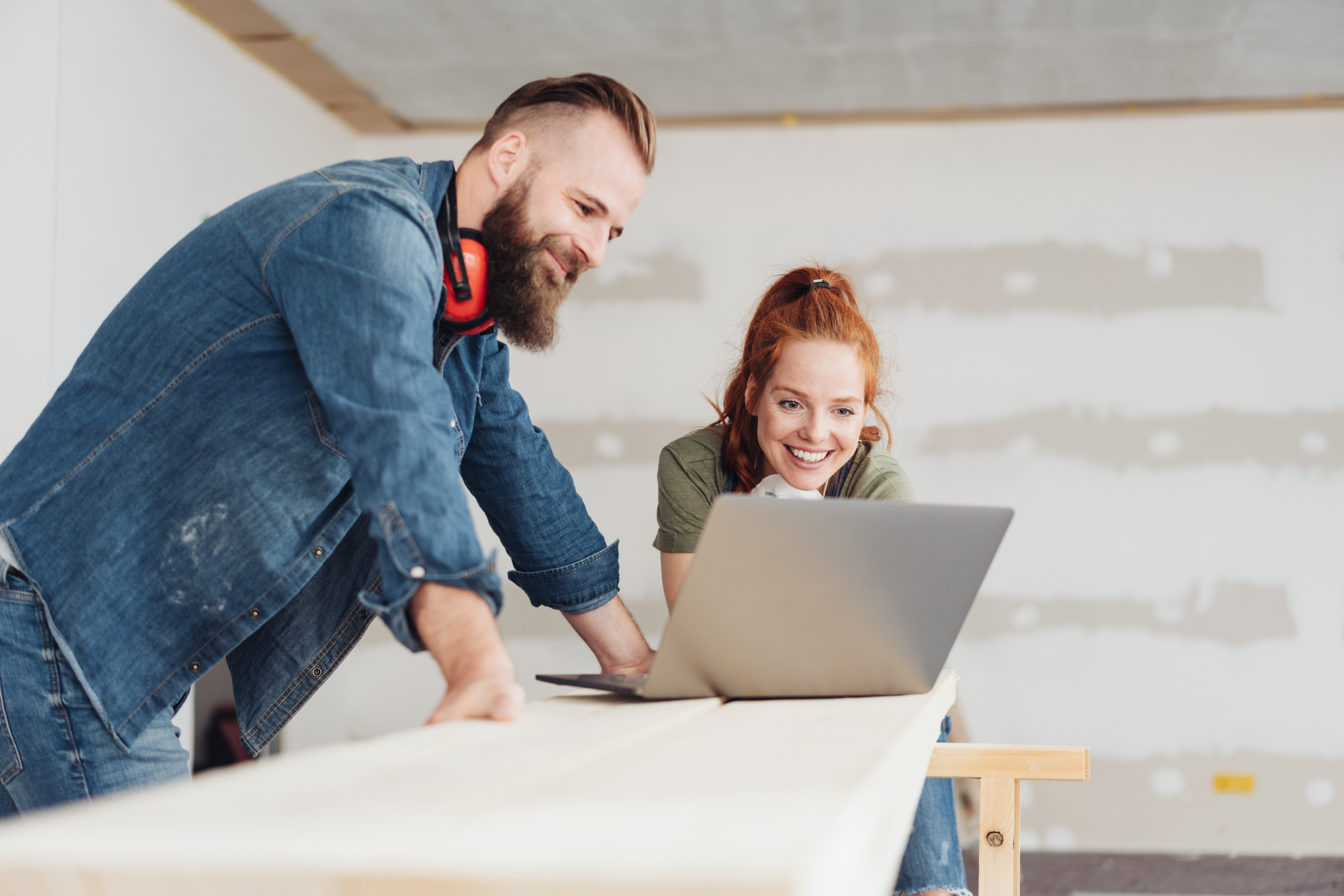 Zwei l&auml;chelnde Personen blicken auf vor sich aufgeklappten Laptop, eine Person tr&auml;gt roten Geh&ouml;rschutz um den Hals, im Hintergrund verschwommen rohverputzte Wand