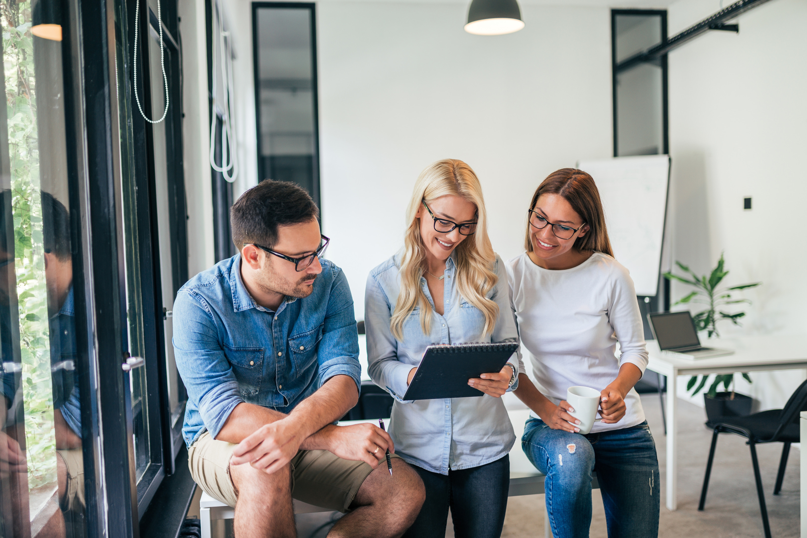 Drei Personen stehen nebeneinander. Die mittlere Person hält einen Notizblock, auf den alle blicken. Im Hintergrund ist ein Whiteboard