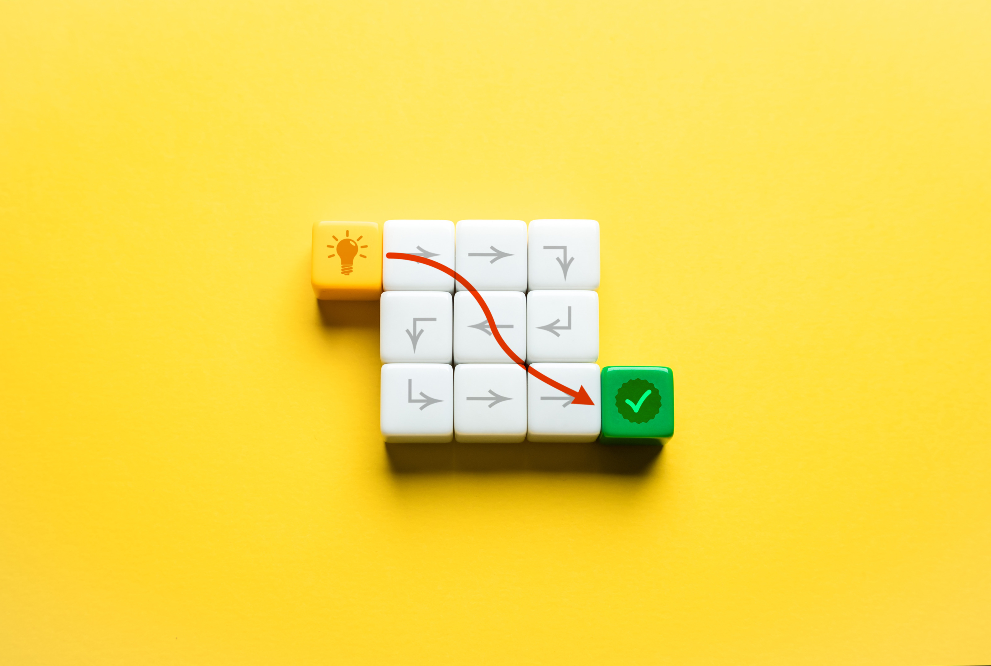 Vor gelbem Hintergrund liegen in der Mitte neun wei&szlig;e W&uuml;rfel. Links oben liegt ein gelber W&uuml;rfel mit einer Gl&uuml;hbirne. Rechts unten liegt ein gr&uuml;ner W&uuml;rfel mit einem Haken. Von der Gl&uuml;hbirne zum gr&uuml;nen Haken f&uuml;hrt eine rote Linie