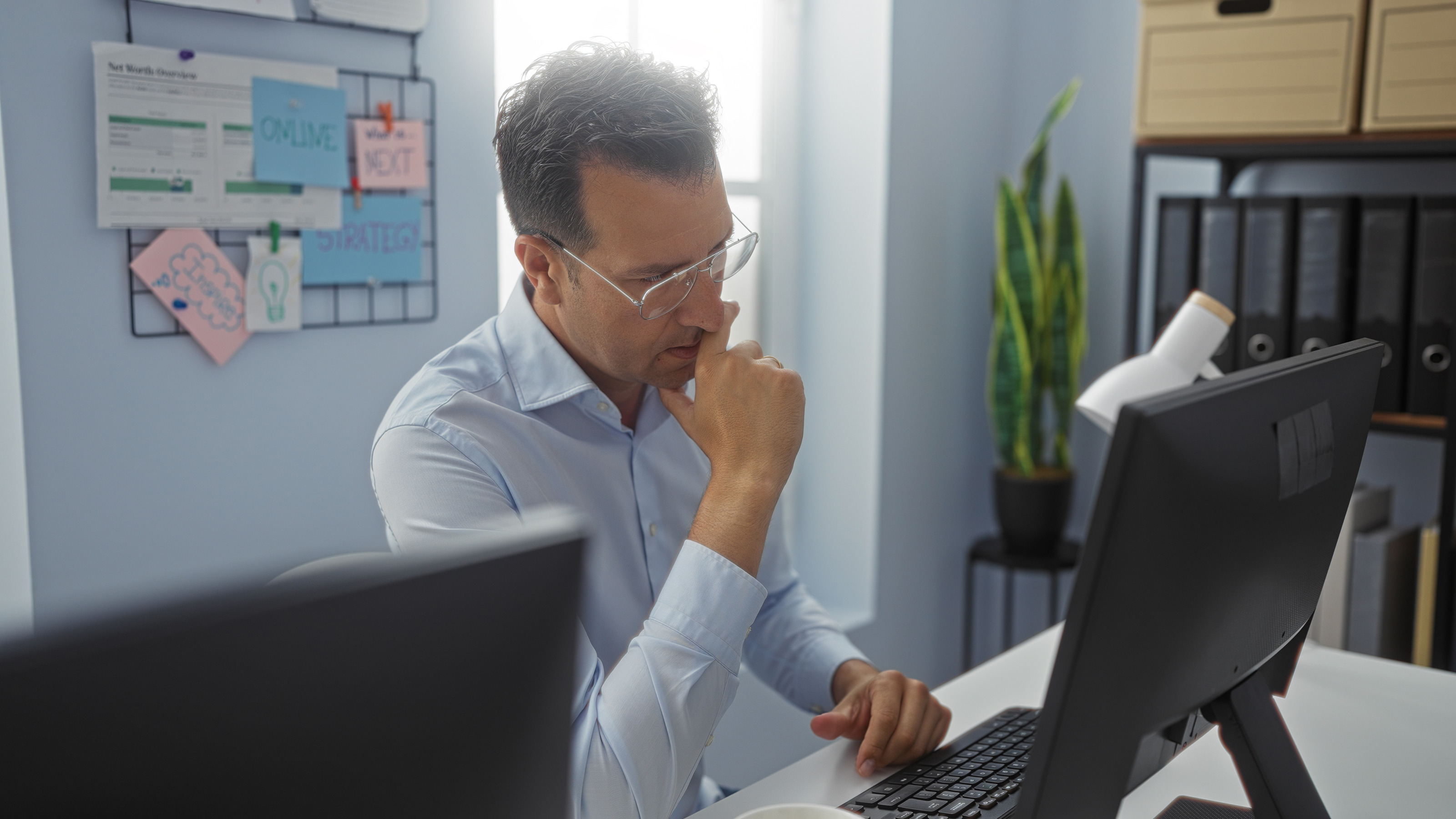 Eine Person mit Brille sitzt an einem Tisch, blickt auf einen Monitor und h&auml;lt eine Hand an den Mund. Hinter der Person h&auml;ngen an einer Vorrichtung verschiedene Zettel mit Stichw&ouml;rtern wie Strategie.