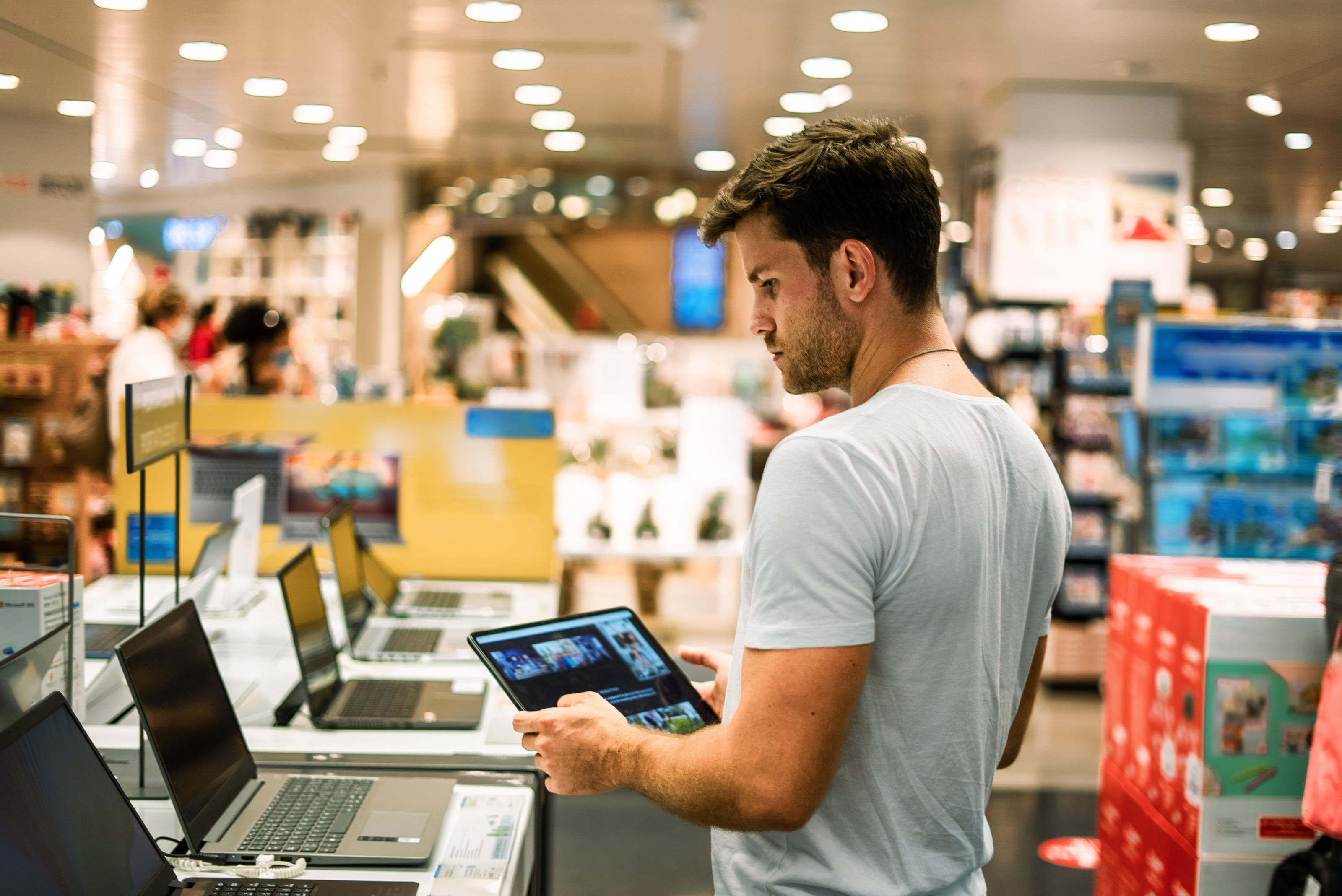 Seitliche Aufnahme einer Person mit kurzen, braunen Haaren, die in einem Elektrogesch&auml;ft neben einem Regal steht und ein Tablet h&auml;lt. Neben der Person stehen mehrere Laptops