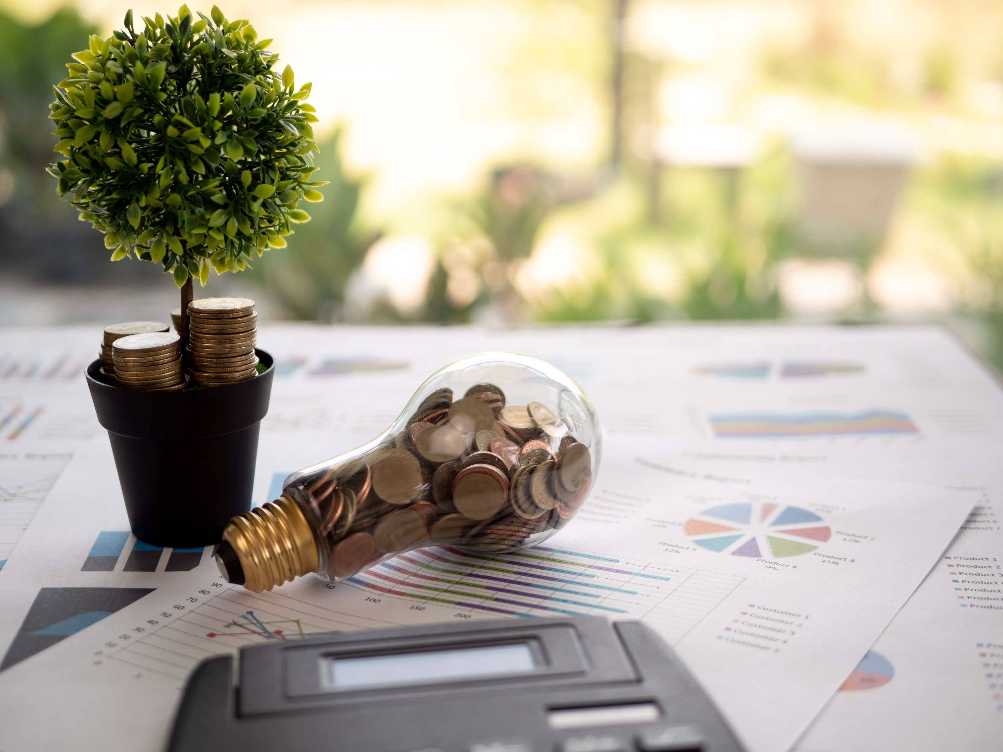 Mehrere Zettel liegen &uuml;bereinander auf einem Tisch. Auf den Zetteln sind verschiedene Statistiken und Diagramme. Auf den Zetteln liegen eine Gl&uuml;hbirne, bef&uuml;llt mit M&uuml;nzen, ein Taschenrechner und eine sehr kleine Topfpflanze, auf der M&uuml;nzen gestapelt sind.