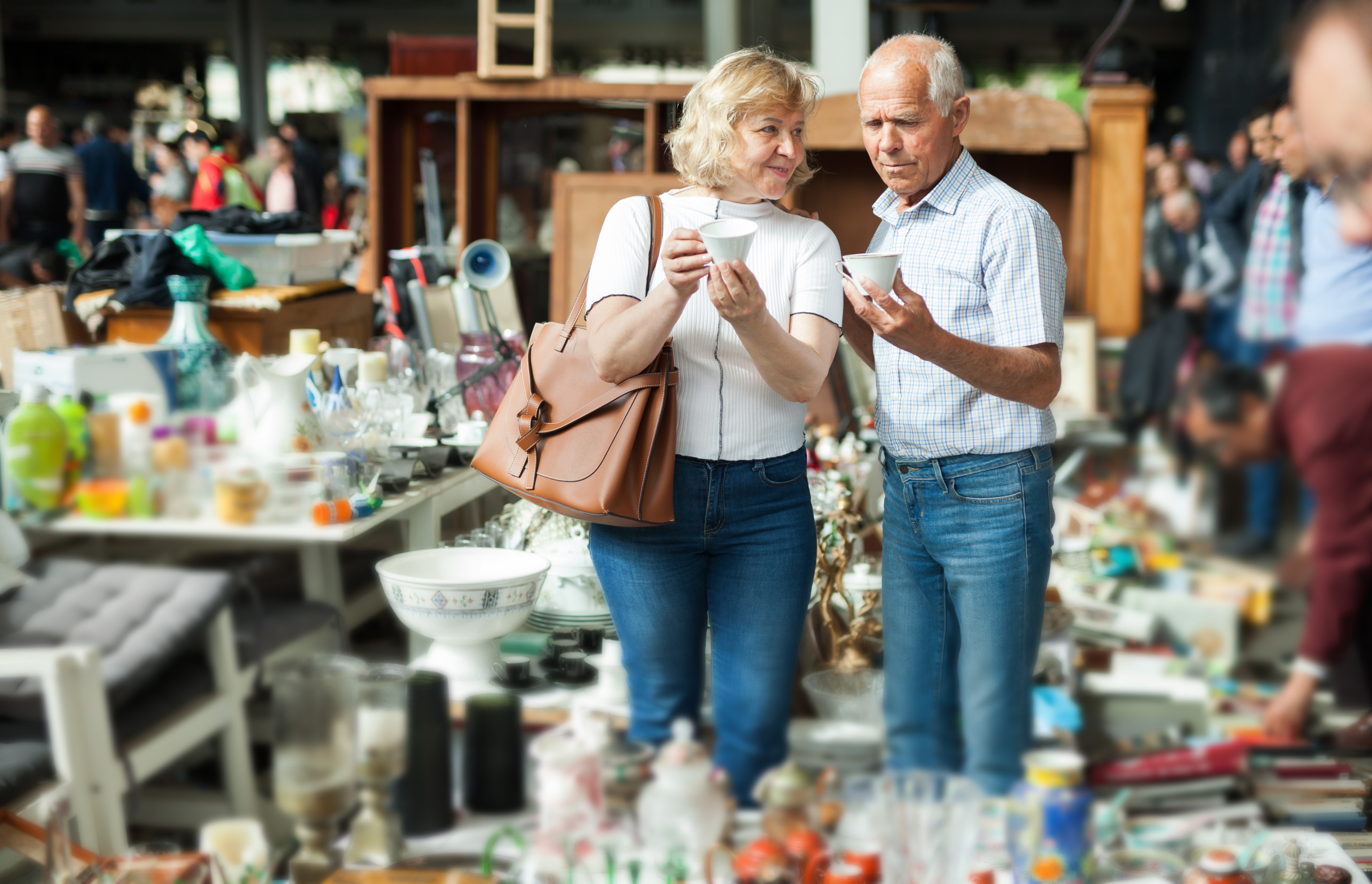 Frontale Aufnahme zweier Personen, die zwischen mehreren Tischen auf einem Flohmarkt stehen. Auf den Tischen sind viele verschiedene Gegenst&auml;nde. Die Personen halten jeweils eine Tasse. Um die Tische stehen viele weitere Personen.
