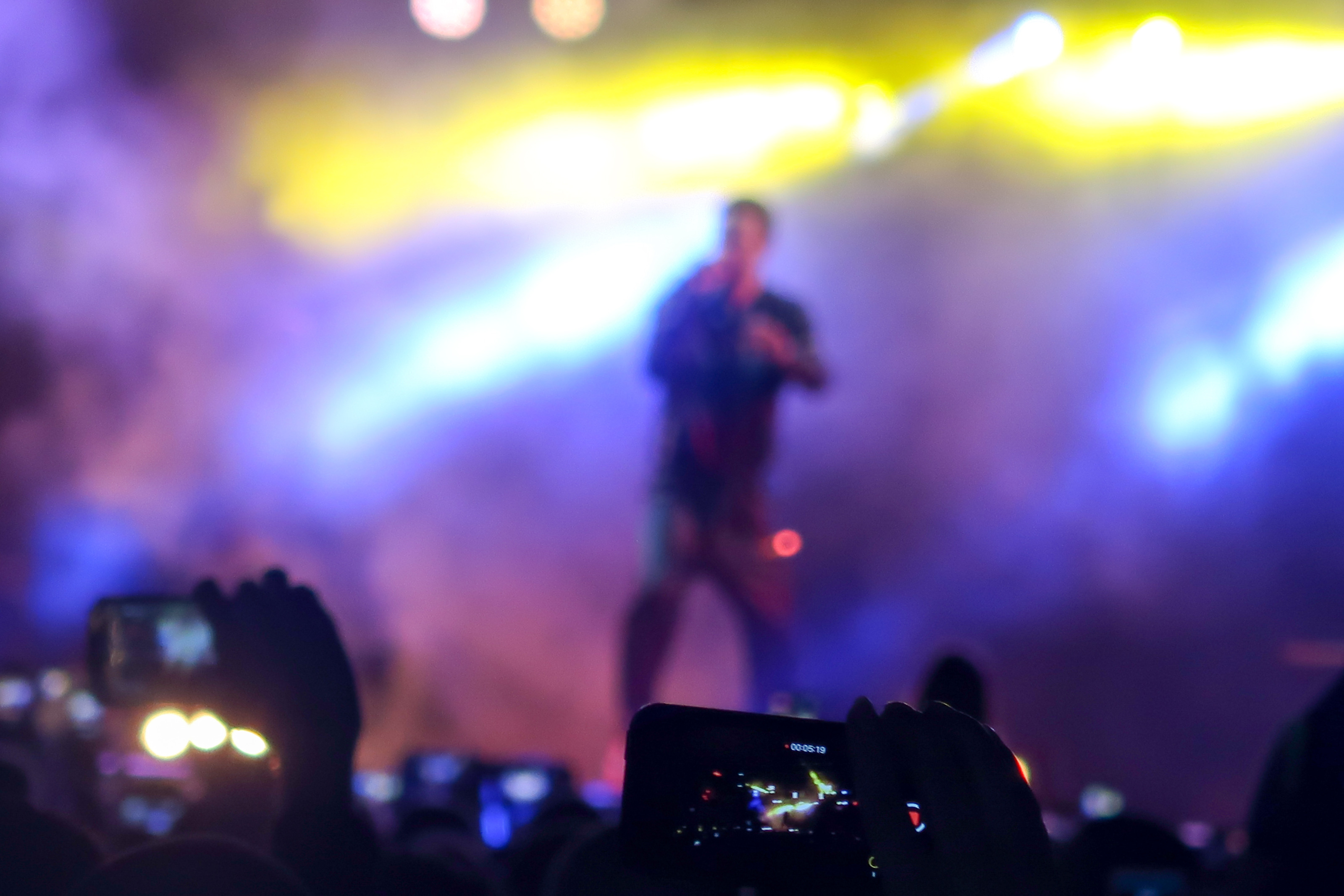 R&uuml;ckenansicht einer Menschenmenge, in der manche Personen ihr Handy horizontal in die H&ouml;he halten und filmen. Ihnen gegen&uuml;ber steht eine Person auf einer B&uuml;hne, die in eine Mikrofon singt