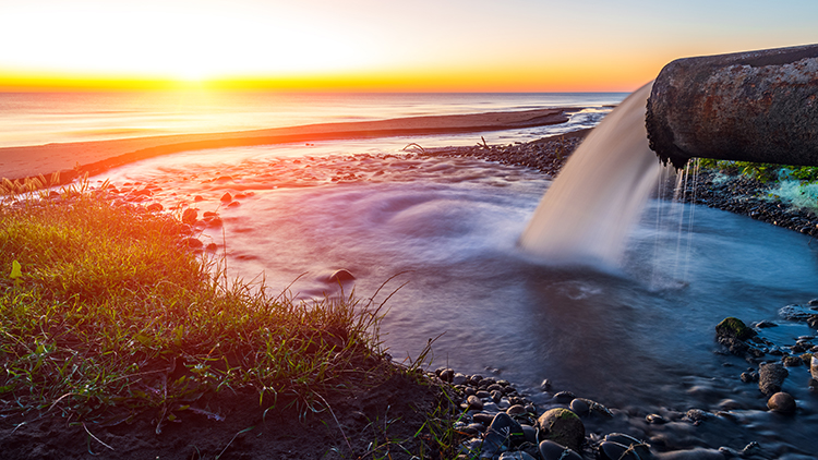 Fl&uuml;ssigkeit rinnt aus Abwasserrohr in ein Gew&auml;sser, im Hintergrund Sonnenuntergang