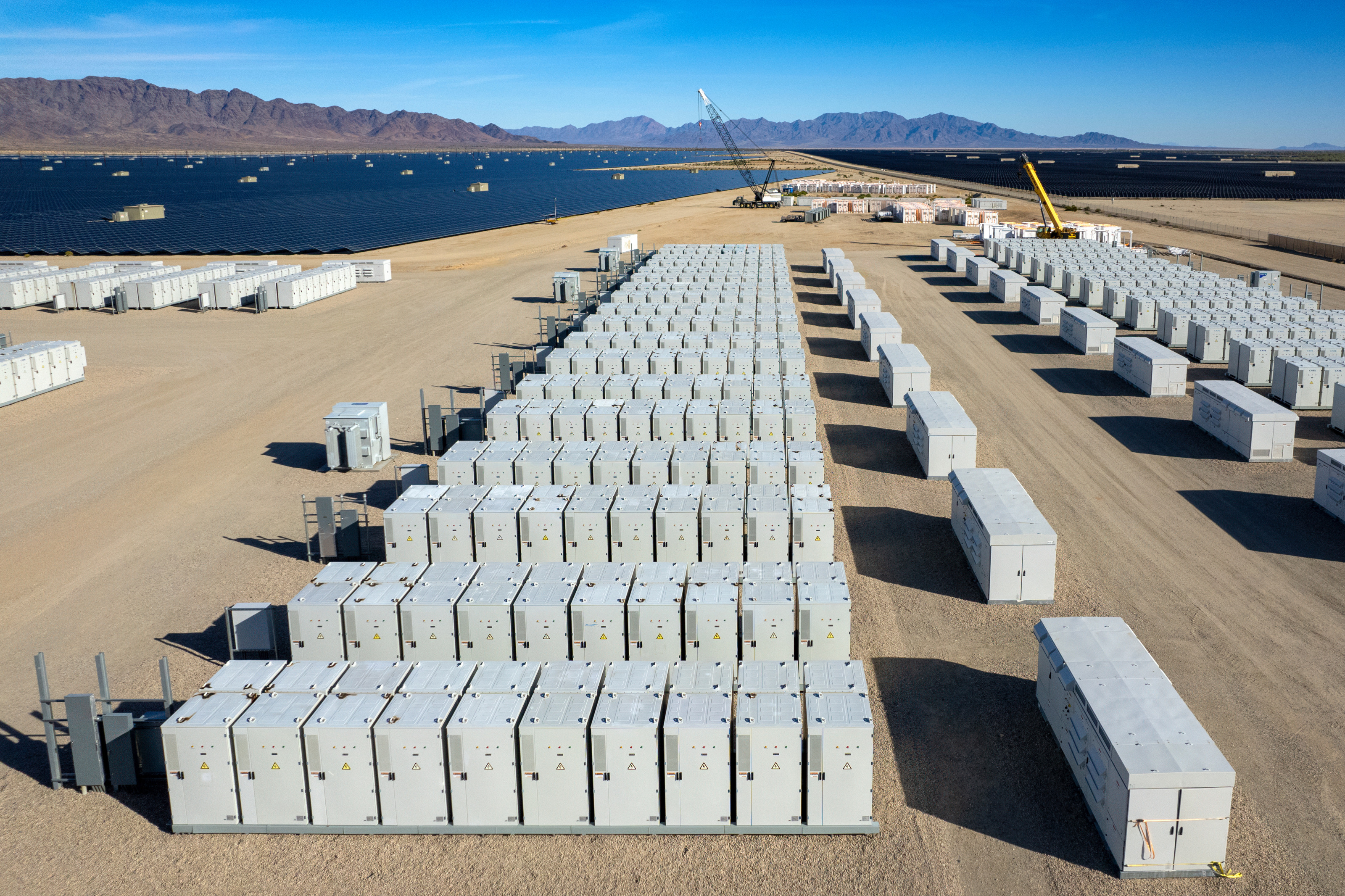 Vogelperspektive auf ein Feld voller Batteriespeicher, im Hintergrund breitfl&auml;chige Solaranlagen und Bergkette unter blauem Himmel