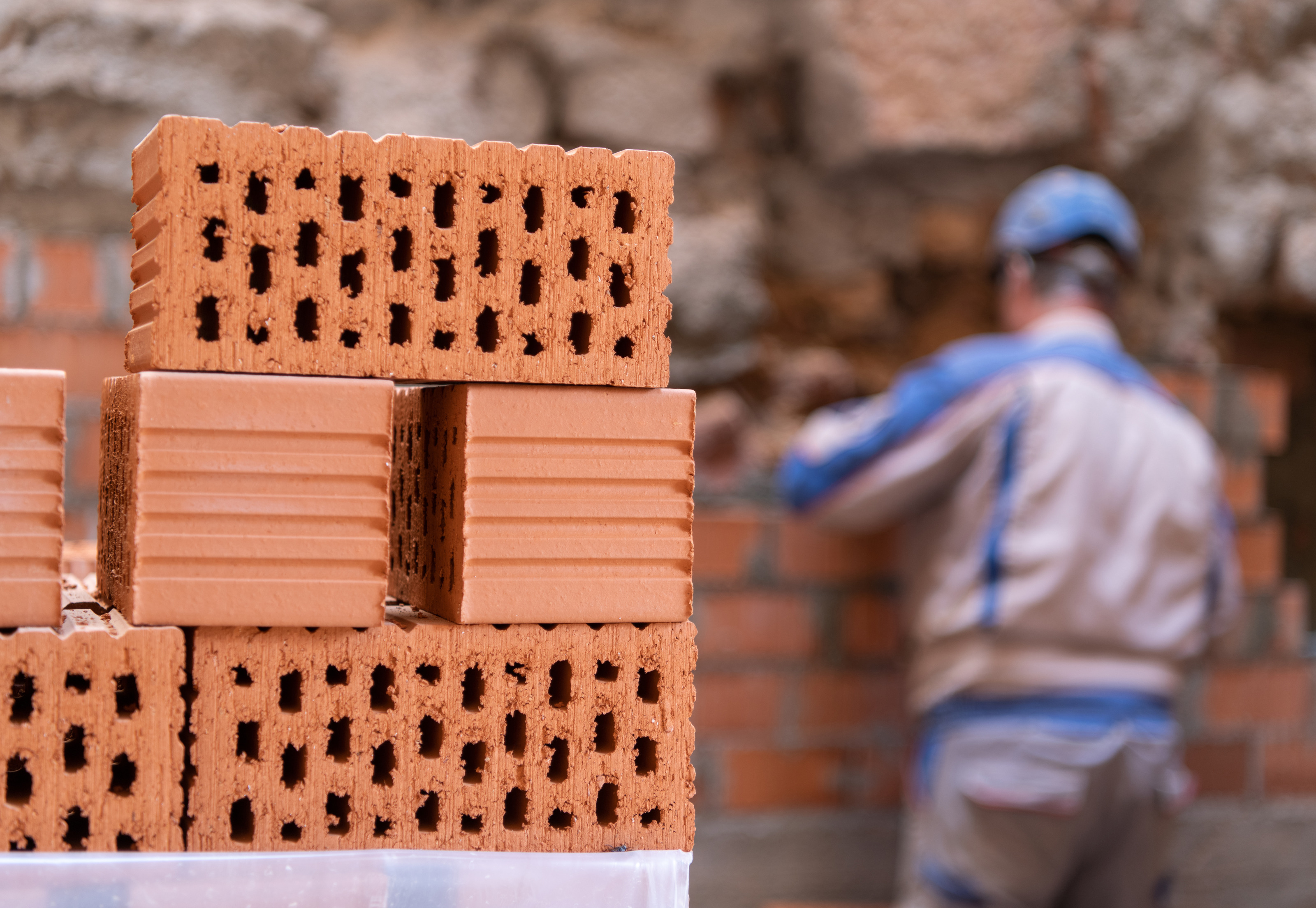 Im Vordergrund sind sechs Ziegelsteine &uuml;bereinander gestapelt. Im Hintergrund ist der R&uuml;cken einer Person mit Helm, die direkt vor einer Mauer aus Ziegeln steht