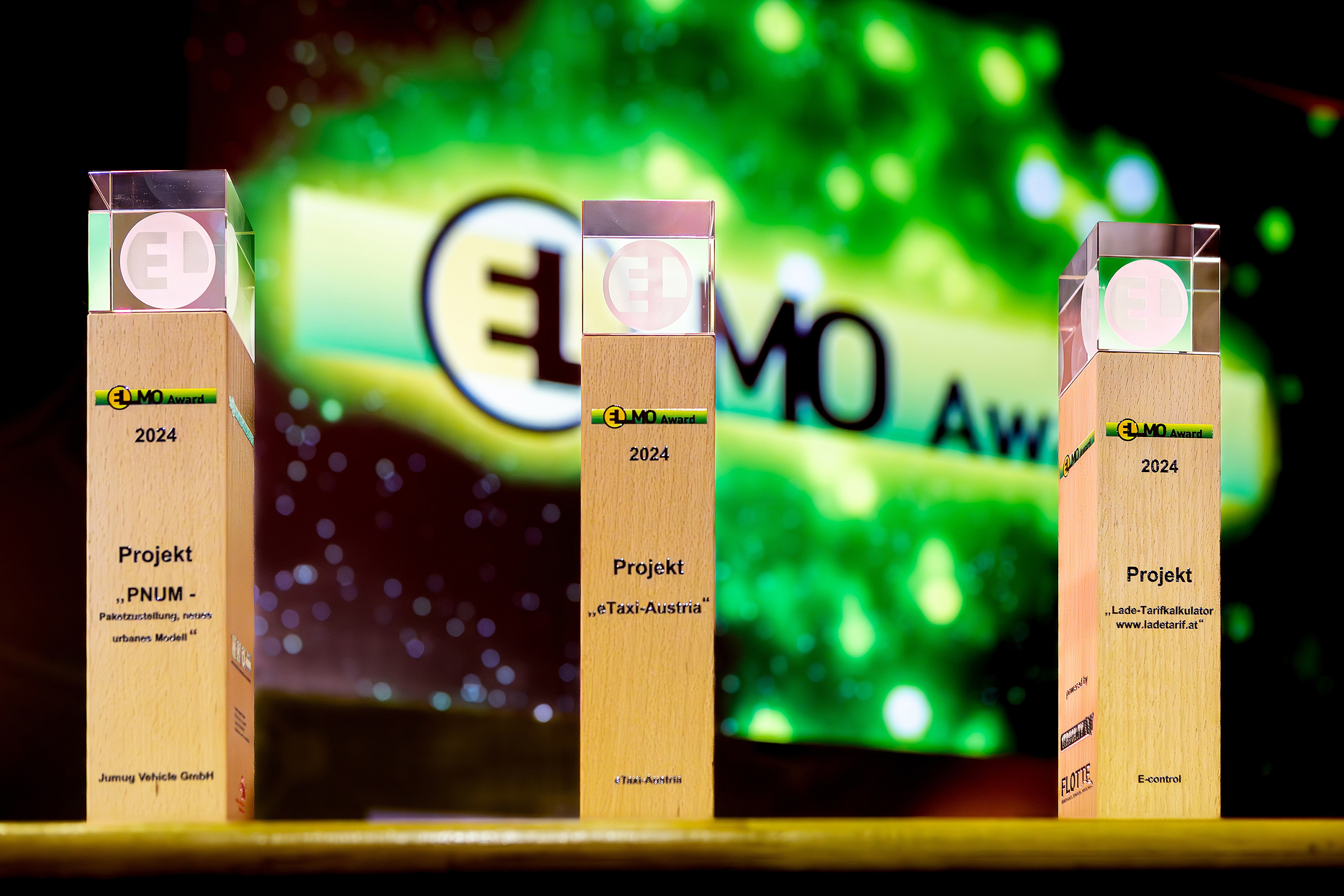 Drei ELMO-Awards nebeneinander stehend: S&auml;ulen aus Holz mit gl&auml;serner Spitze und der Aufschrift Elmo-Award 2024 Projekt, im Hintergrund das Elmo-Logo auf gr&uuml;nem Licht