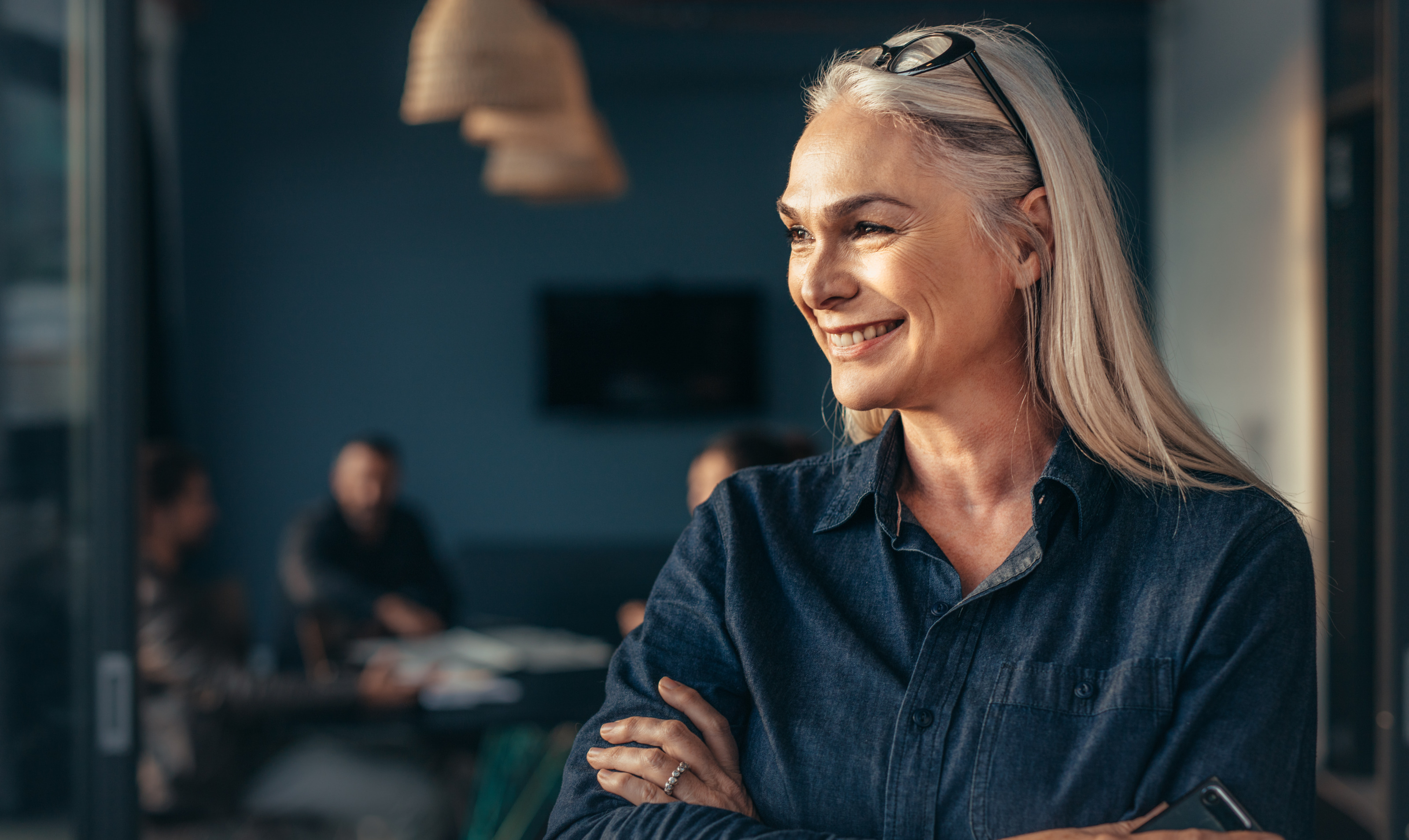 Porträt einer Person mit langen blonden Haaren trägt eine Brille in den Haaren und eine blaue Jeansbluse während sie freudig auf die Seite blickt, im dunklen Hintergrund sitzen Personen an einem Tisch