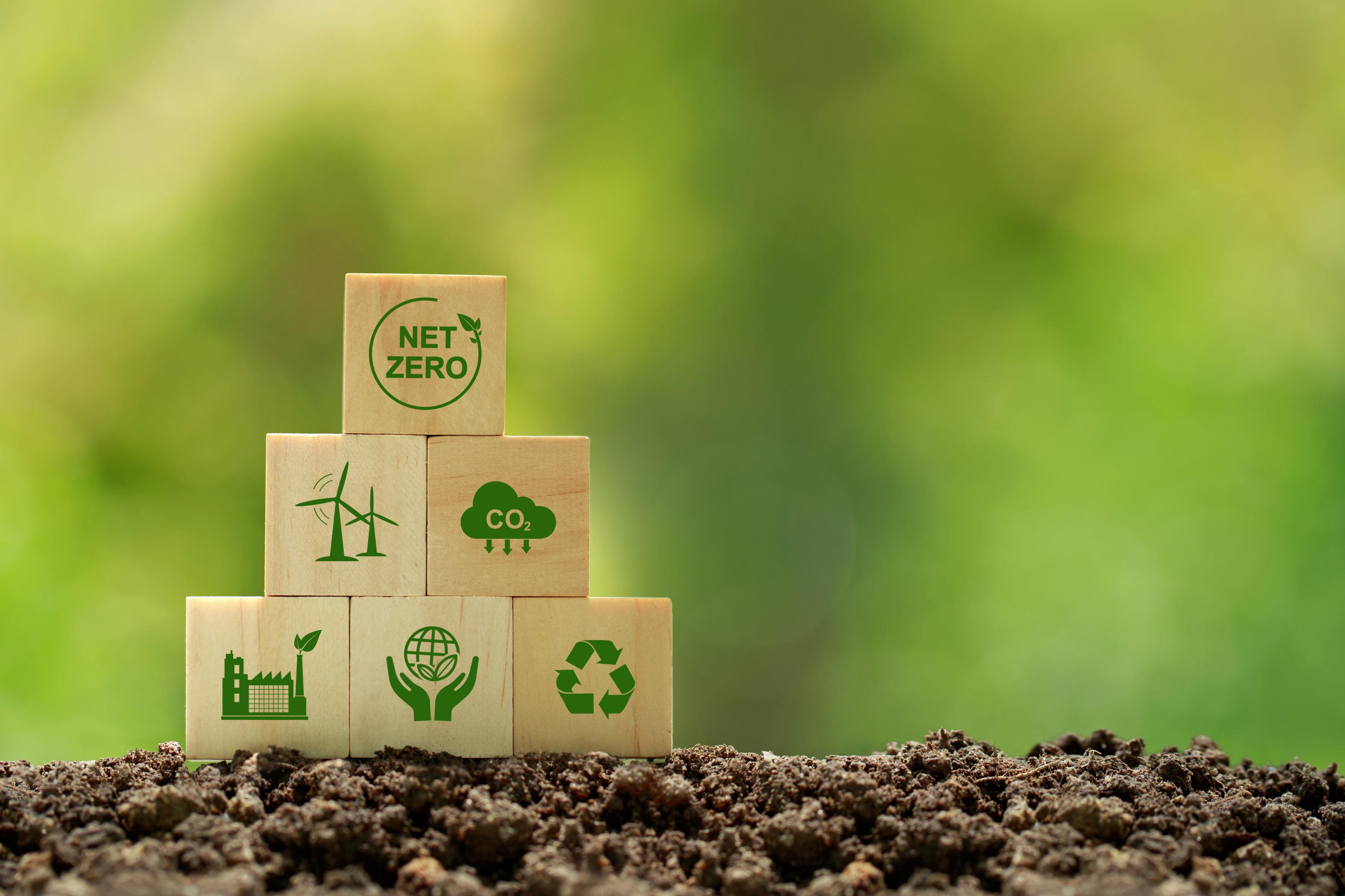 Mehrere Holzw&uuml;rfel aufeinander gestapelt auf Erde mit gr&uuml;nen Symbolen zum Thema Klimaschutz, oberster W&uuml;rfel mit Schriftzug Net Zero, Hintergrund verschwommen gr&uuml;n 