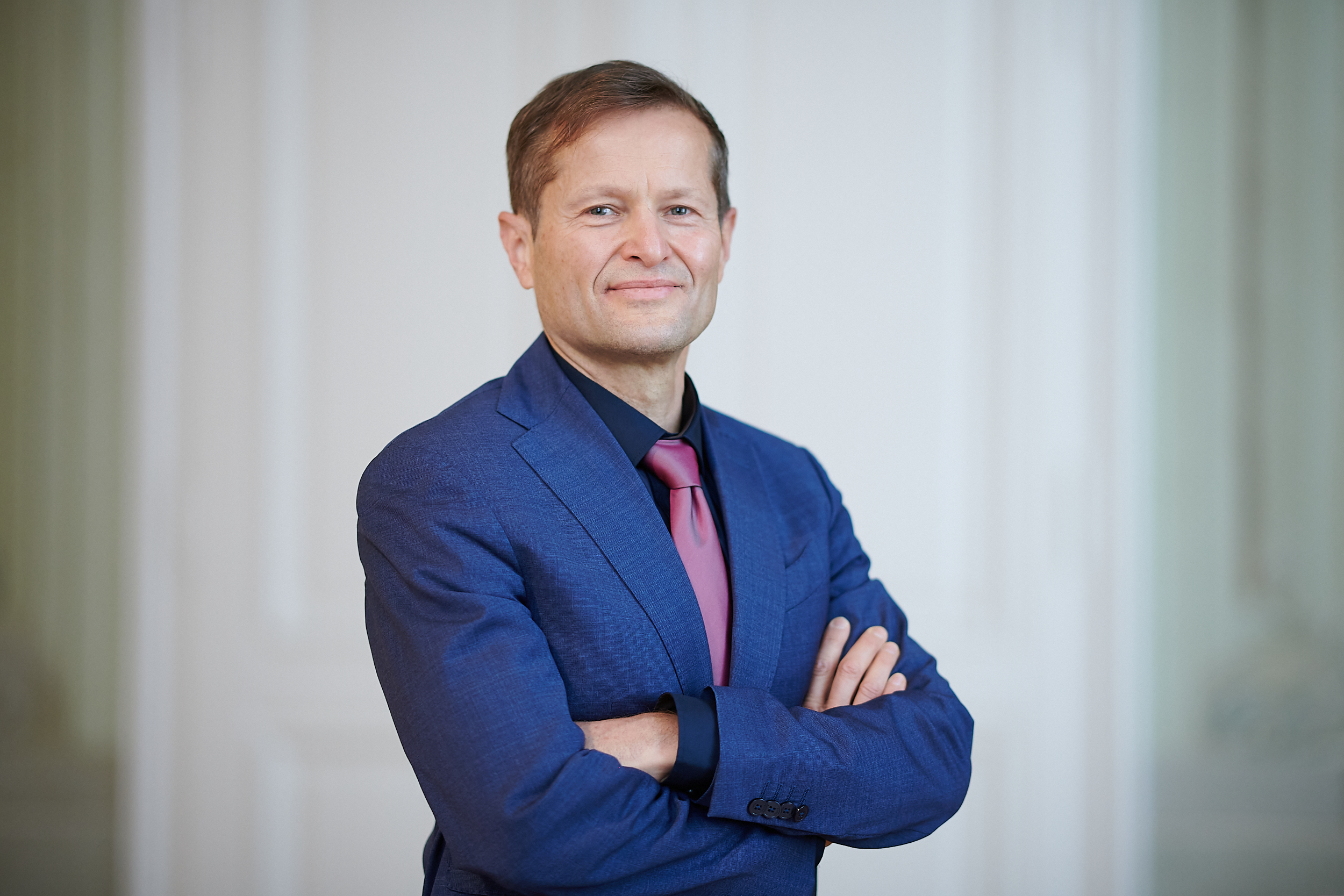 Portrait einer sanft lächelnden Person in dunkelblauem Anzug mit Armen verschränkt vor Körper vor verschwommenem Hintergrund