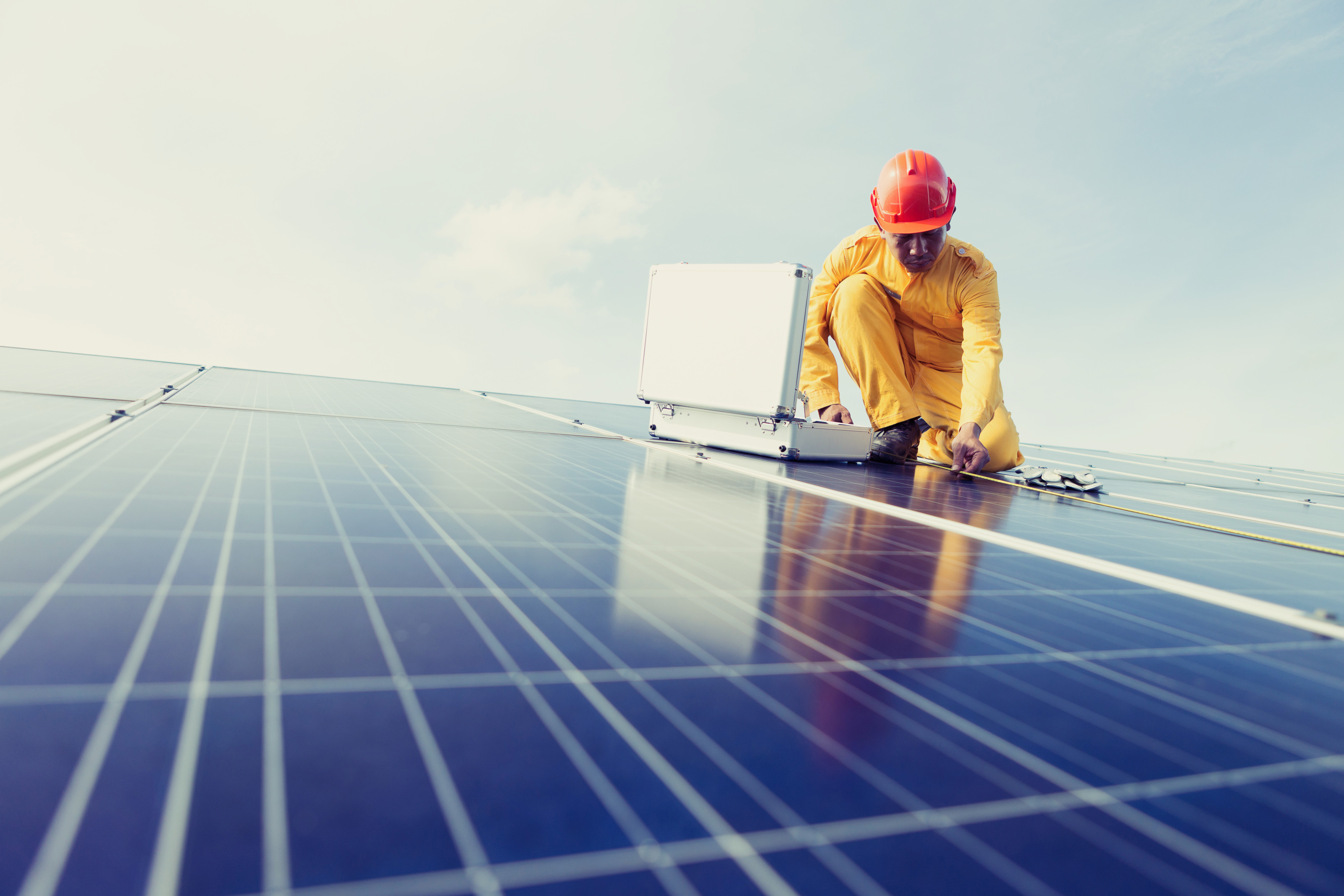 Person in gelbem Arbeitsoverall mit orangem Helm kniet auf Solarpanelen und misst mit Ma&szlig;band, vor Person ge&ouml;ffneter Koffer 