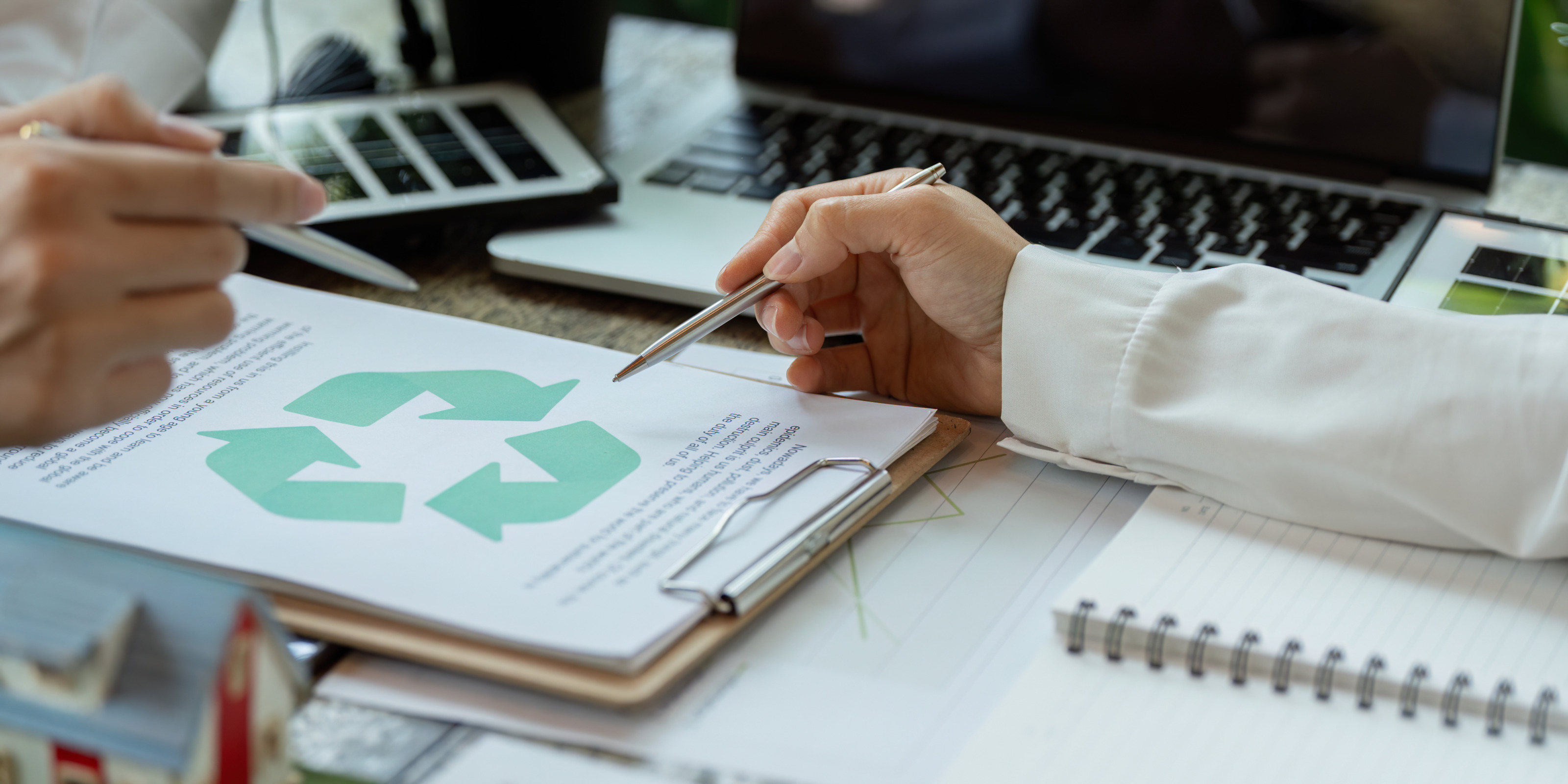 Auf einem Tisch liegt ein Klemmbrett mit einem Zettel, auf dem das Recyclingsymbol ist. Zwei Personen deuten jeweils mit einem Stift auf das Symbol