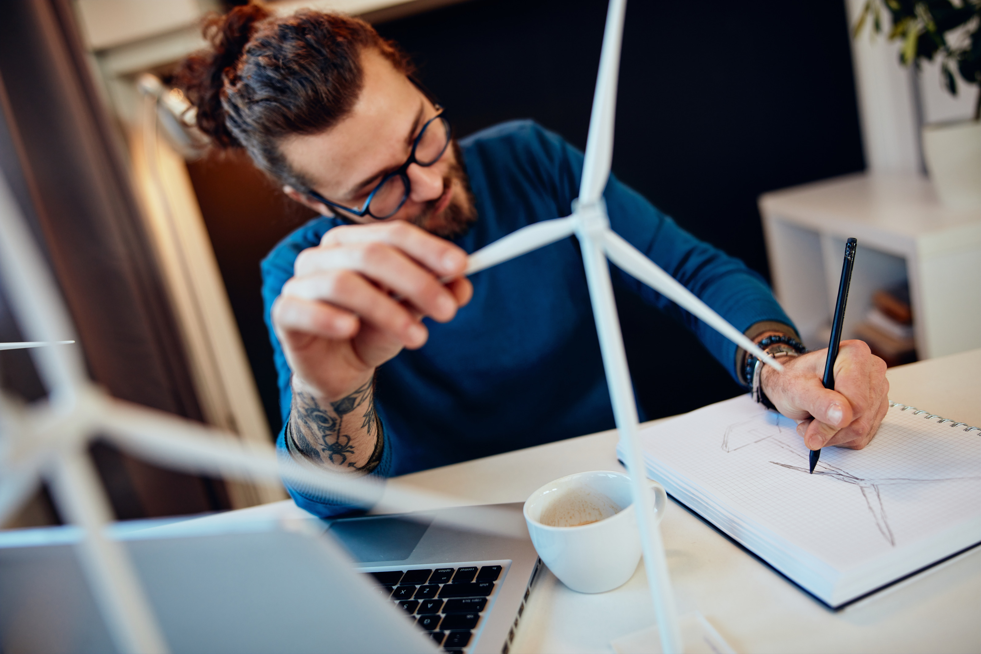 Person mit Brillen sitzt an Schreibtisch, skizziert Windrad auf Block und h&auml;lt in einer Hand Modell eines Windrades, am Tisch aufgeklappter Laptop und Kaffeetasse