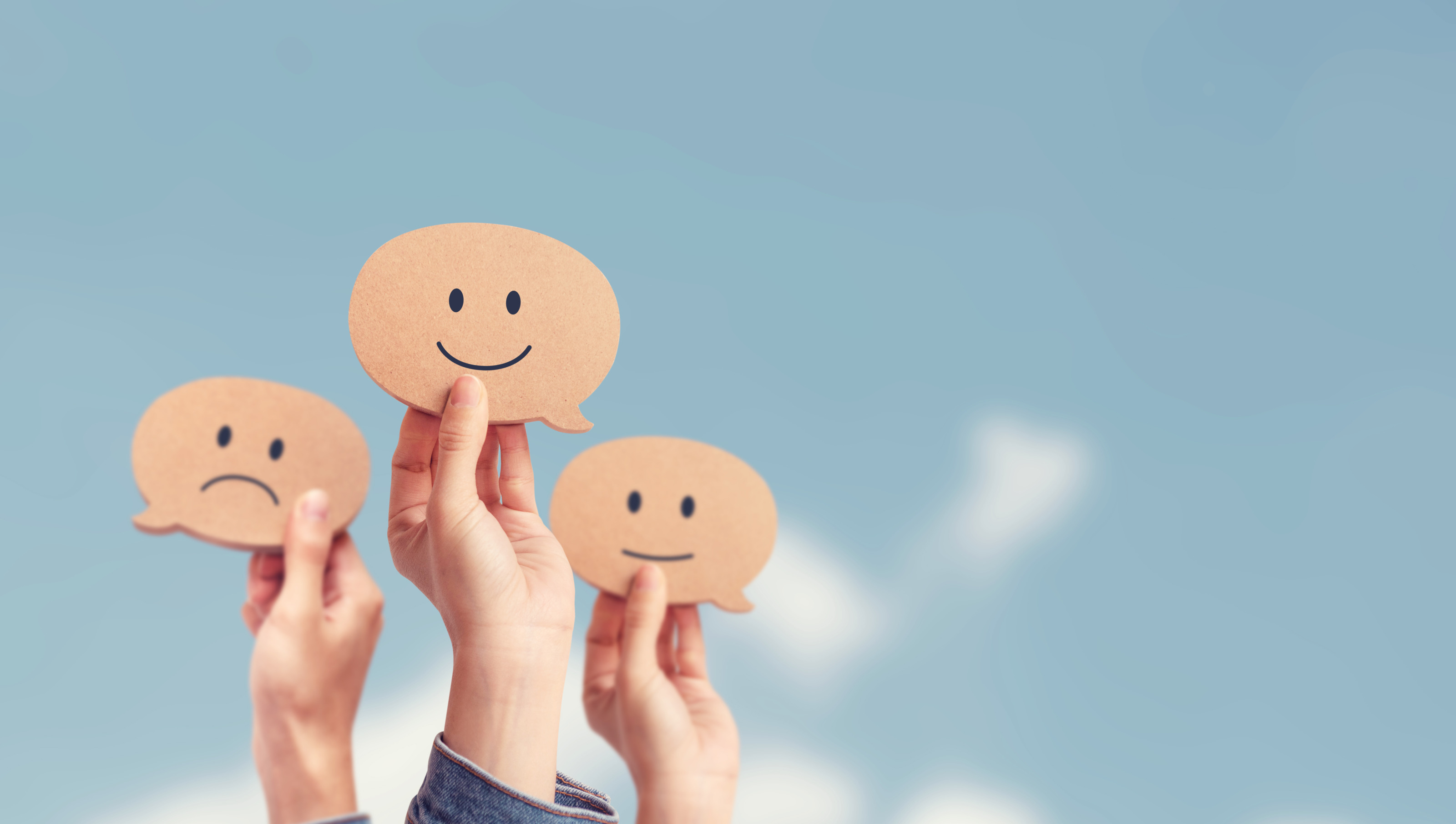 Vor blauem Himmel sind drei H&auml;nde in die Luft gestreckt. Jede Hand h&auml;lt ein Smiley. Ein Smiley l&auml;chelt, eines blickt neutral und das dritte zieht die Mundwinkel nach unten